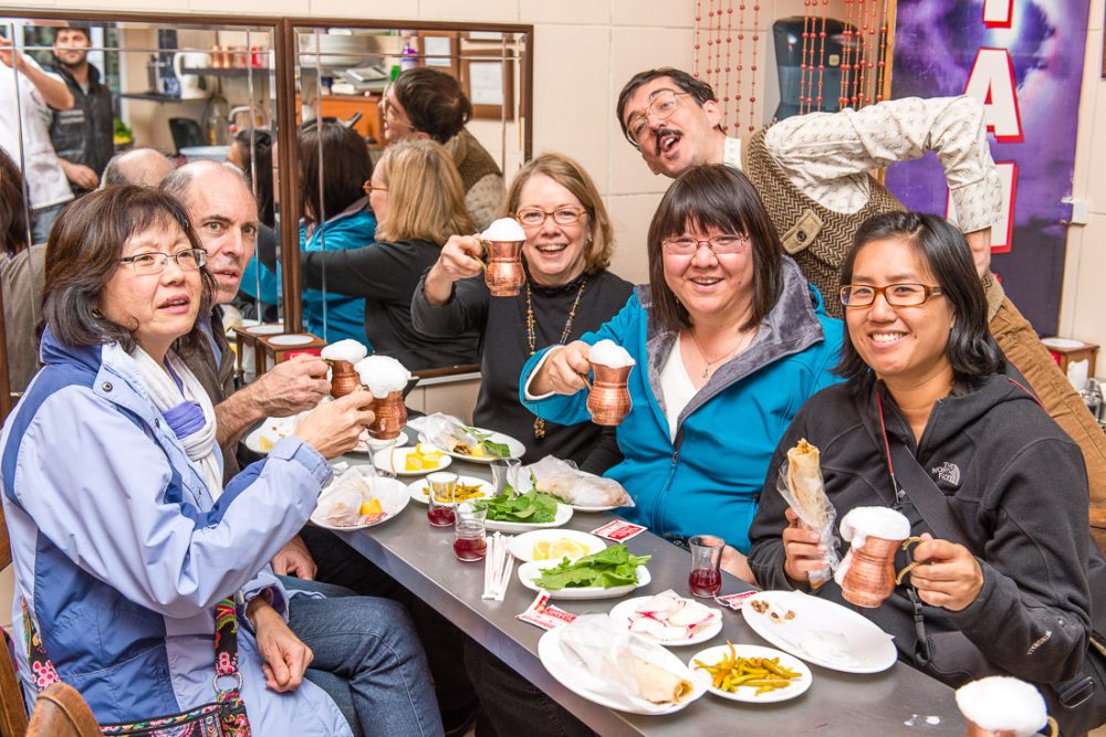 Women traveling solo: A group of 5 people sitting at a table (and one standing behind). The plates on the table are mostly empty. The people raise mugs of something that has white foam overflowing the top.