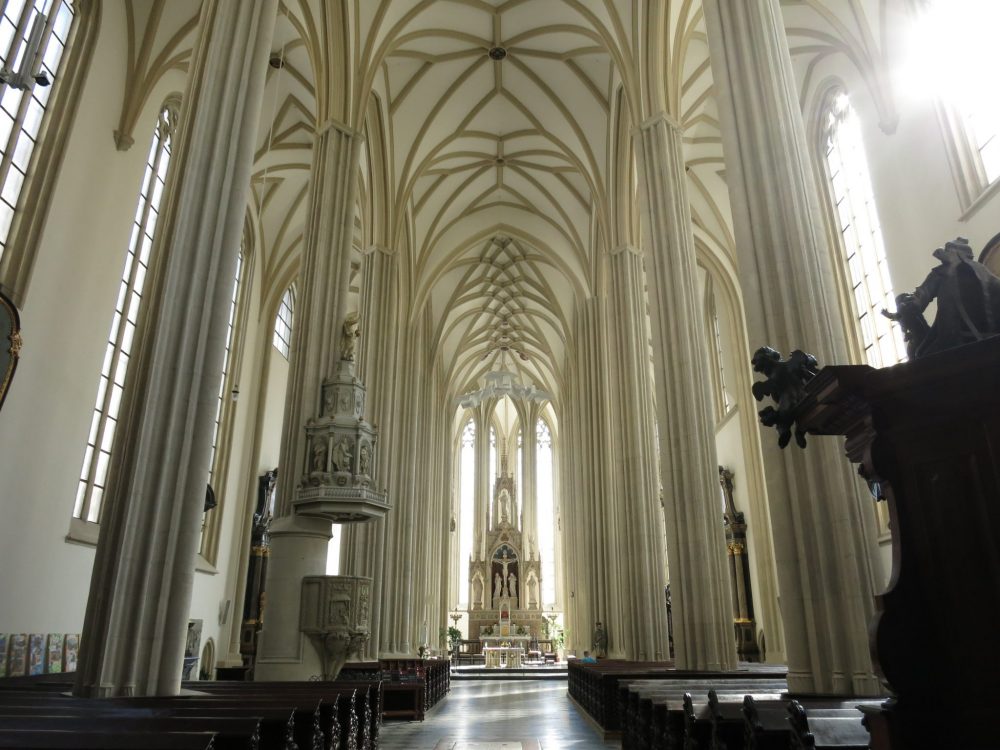 A tall central aisle with white columns on either side and gothic ceiling overhead. Far ahead is an ornate altar. Macabre sightseeing in Brno