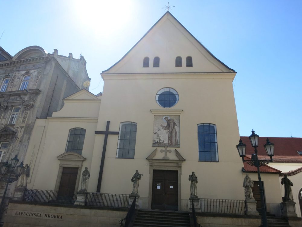 A simple church building with a flat front, a door in the center and two high windows on either side. Above the door is a fresco or mozaic of a saint? Or Jesus? giving a blessing. Above that, a small round window. Along street leve is a line of 6 statues.