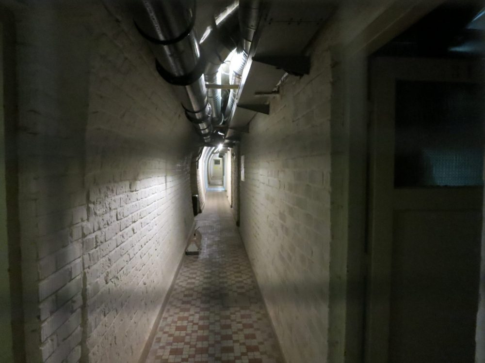 Looking straight down a long, very narrow tunnel. The floor is tiled anad the walls are white-painted brick. Above, a wide pipe extends along the tunnel. In the far distance is a door.