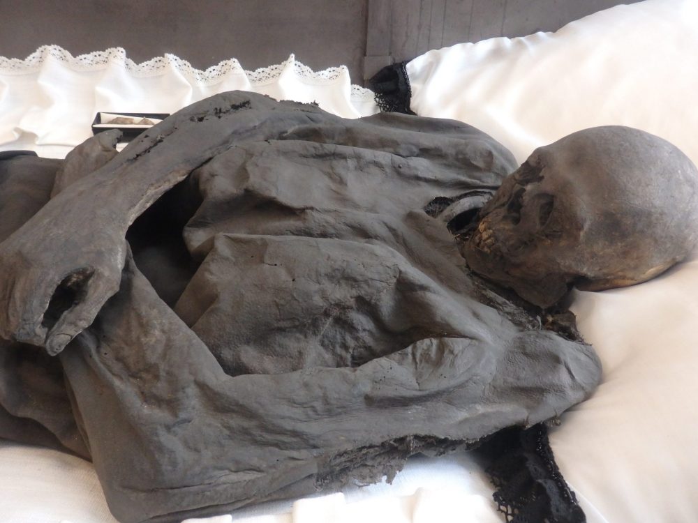 The top half of the body is visible in the photo. Lying on white cloth, the clothing and the arms and hand, as well as the skull are all the same dark grey color. The arms are crossed over his waist.