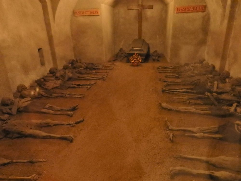 A dimly lit, square room. At the far end, straight ahead, is a simple coffin with a cross on the wall above it. On both sides of the room are the bodies, lying with their heads to the left or the right wall, bare feet toward the center. They are shriveled and skeletal, but have clothing and skin.Macabre sightseeing in Brno.