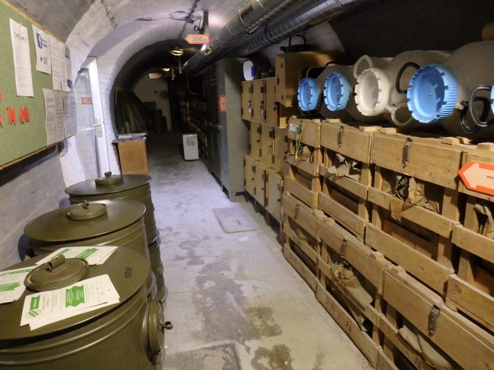 Looking down a wider tunnel this time. ON the left, a bulletin board on the wall with some cylindrical containers standing upright underneath it. On the right, a long pile of crates, with more smaller cylindrical containers lying on their sides on top of the crates.