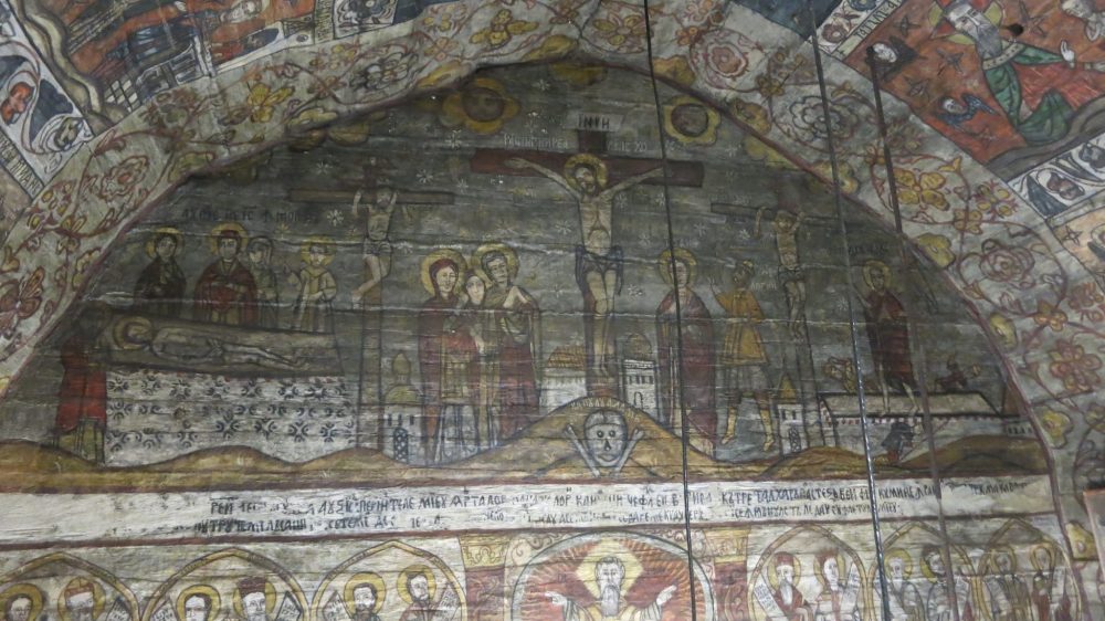 The image is semi-circular under the arch of the roof (also painted). Jesus in the center on the cross, the two thieves on either side on smaller crosses, and various figures with haloes among the crosses on the ground below them. Very primitive images, fading.