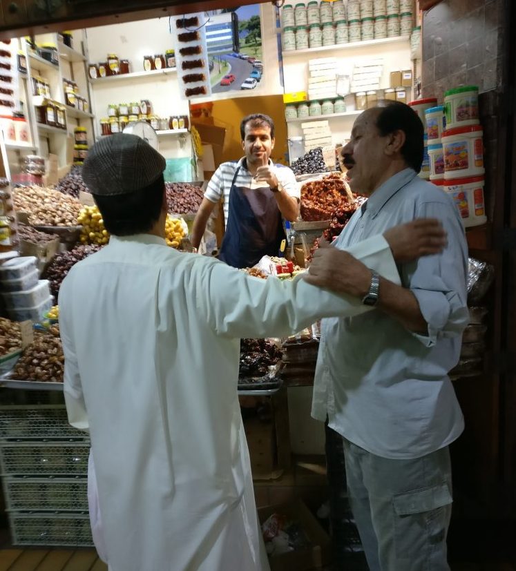 Market trader surrounded by bins of dried fruits and nuts facing the camera giving the thumbs up. Two men greet each other in front of the stall.