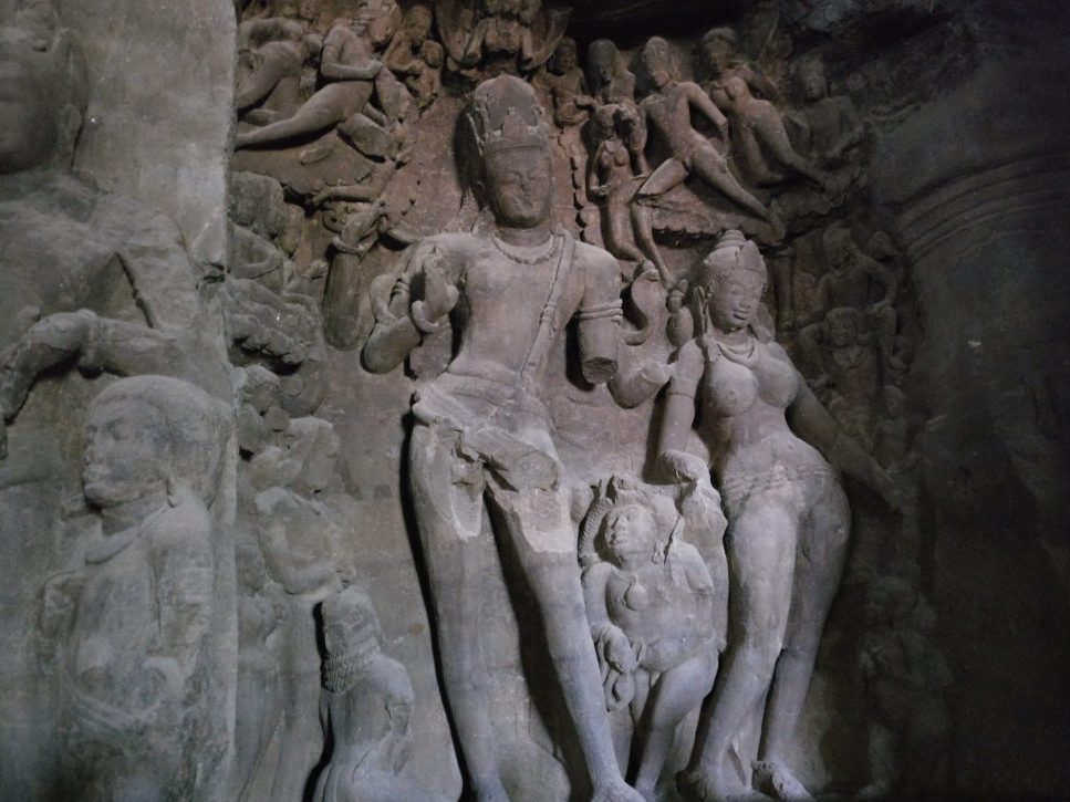 The stone-carved bas-relief has a standing figure in the center with a smaller female standing figure to its right. Around both of them are a multitude of other much smaller human figures. Elephanta Island travel guide