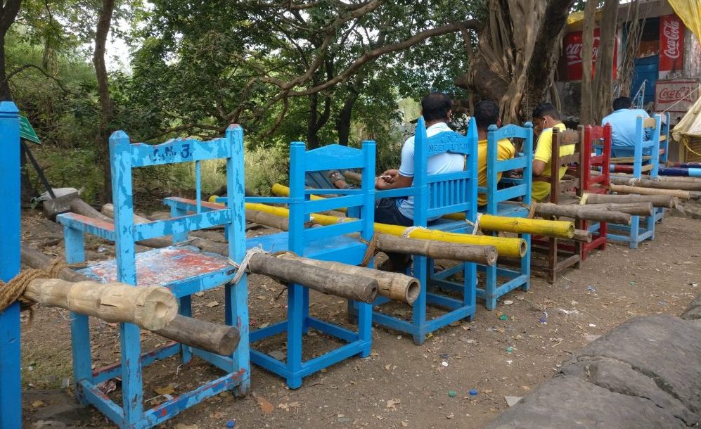 A row of bright blue wooden chairs stand with their back to the camera. EAch has two bamboo poles lashed to it with prope, one on either side of the seat. Men sit on some of the chairs. Elephanta Island travel guide.
