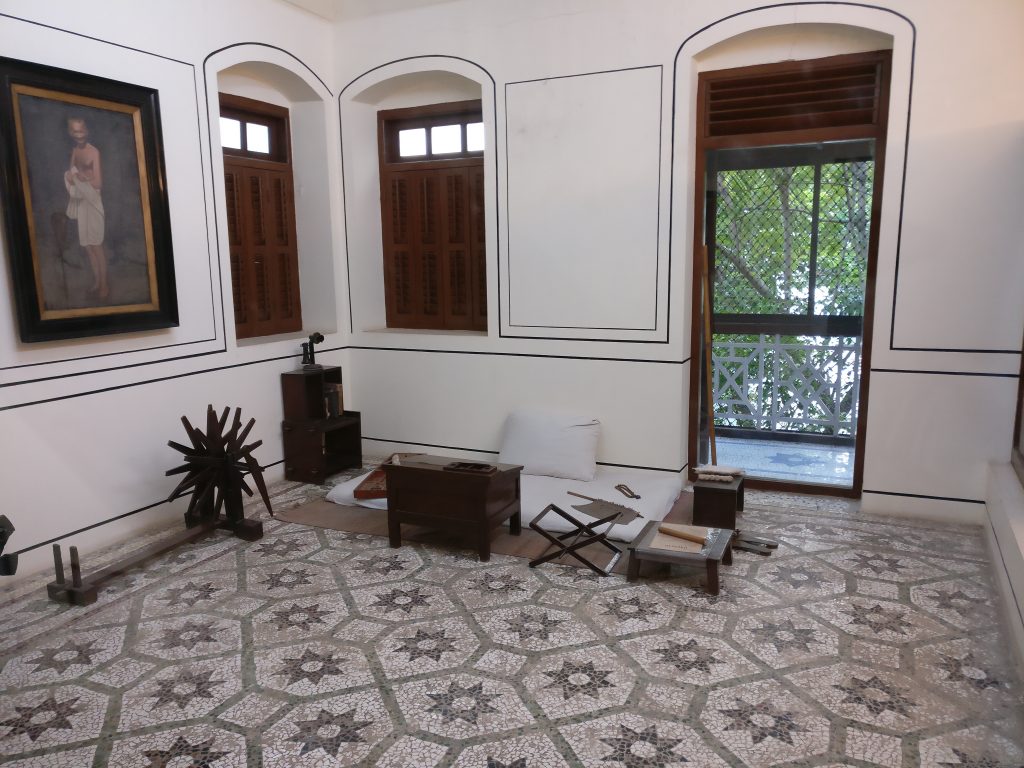 A room with a tile floor, almost empty except for a mattress and small table and writing tools. A painting on the wall of Gandhi.