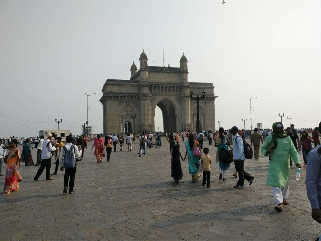 An archway as seen across a plaza with many people walking in different directions. The archway has 3 big arches, the largest in the middle.