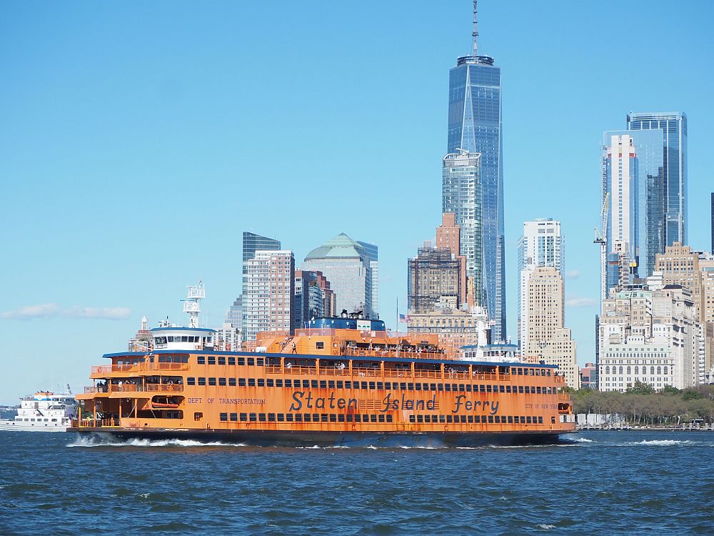 The Staten Island crossing dark blue water. The ship is a dirty orange color with a black hull. The sides are lined with square windows and at the end are some open spaces on each floor with railings at their edges. Behind the ship is part of the Manhattan skyline: a cluster of tall buildings. The Staten Island Ferry is a great way to see New York City on a budget.