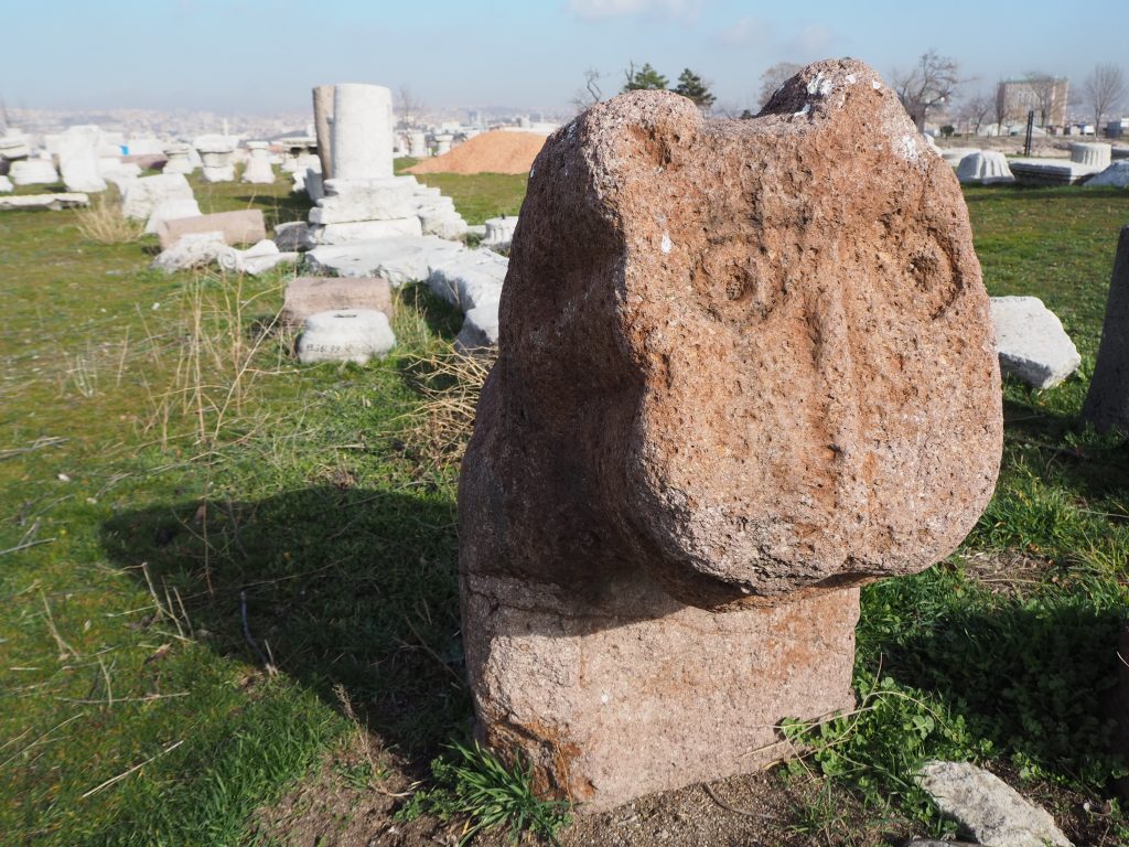 A reddish stone figure with a face of an animal, looking like a Moomin. Behind are various bits of buildings in white stone.