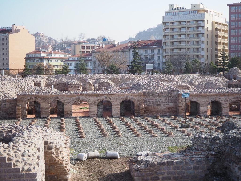 A low flat section with even rows of bricks piled into low columns. Behind that, a row of arches, and modern buildings beyond that.