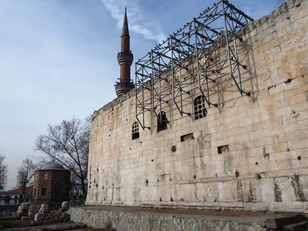 A large flat white stone wall, with some metal scaffolding in one section, and a minaret behind it.