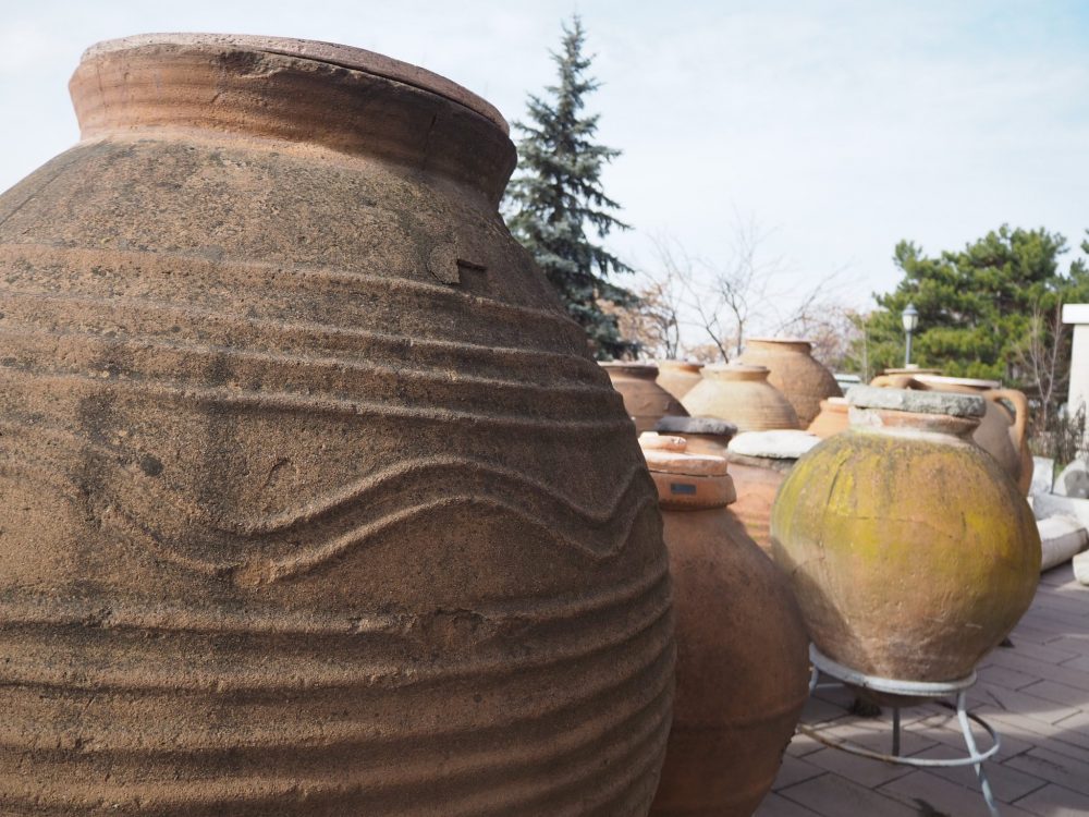 The pots are clay and quite large. The nearest one has simple lines in relief around the outside: some are wavy but most are straight horizontal lines. The pots behind are similar but many are not decorated at all. One in the photo has at least one handle on its side.