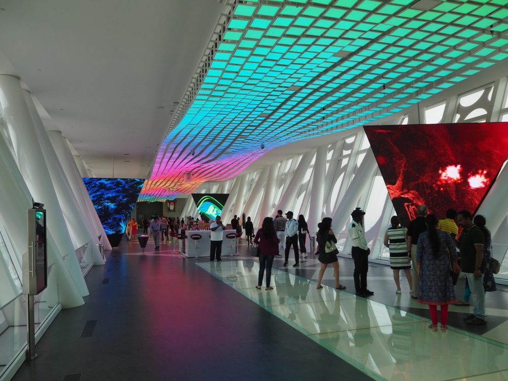 A long room, with a scattering of people. The walls on either side lean outwards at the top, with big, white columns that also lean outwards, tilted left or right. The floor is dark, except for the greenish-gray glass panels down the center. The ceiling looks like it has a strip of tiling down the center, but the tiles are lit up in blues and pinks, and the whole thing is wavy, rather than flat.