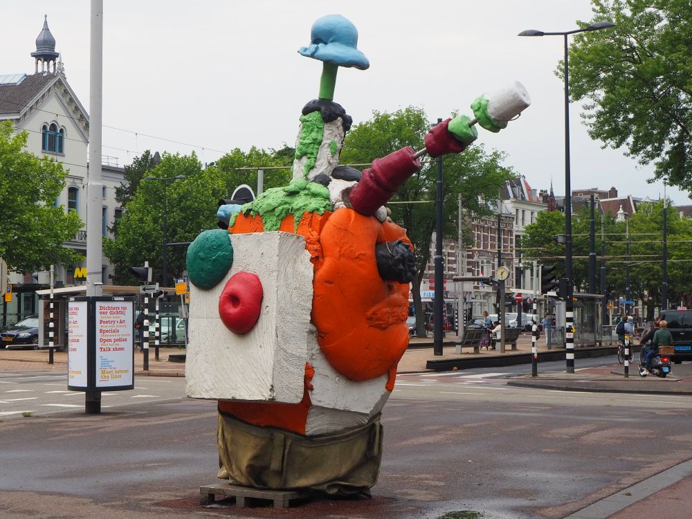 Hard to describe this statue. It seems to be an assemblage of odds and ends: pieces of concrete, some sort of bright orange plastic or foam. It has two shapes sticking out of it: one on the side has some sort of red objects skewered on it and something that looks like a giant marshmellow on the end. The shape sticking out on top has a blob of blue on the end that is shaped something like a hat.