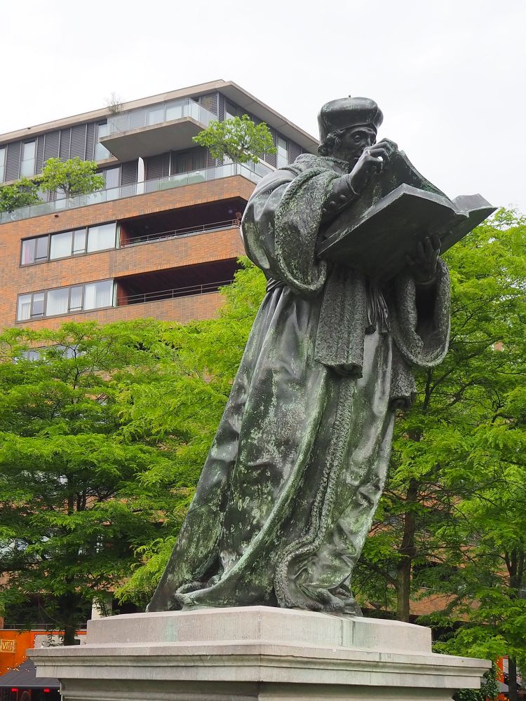 bronze full-size statue of Erasmus in robes, wearing a beret-like hat. He holds a large book and turns the page as he reads.