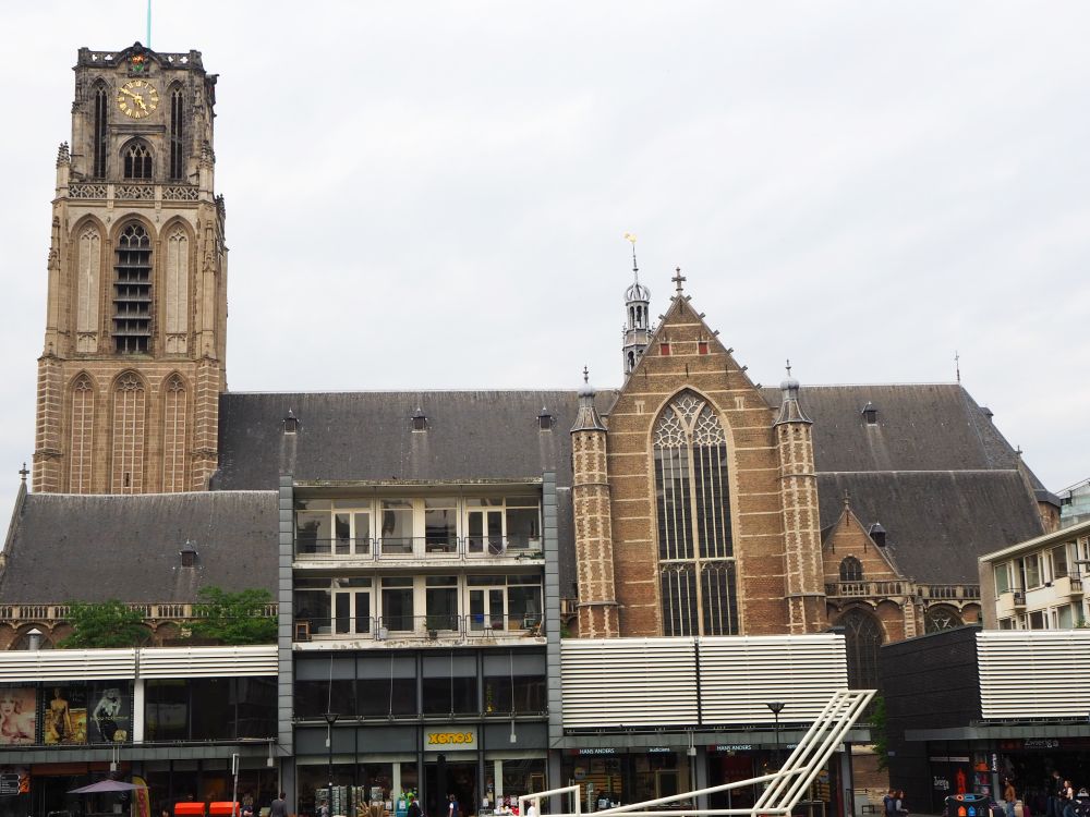 The church is seen from the side, with the church tower on the left. About halfway down the side, a large gothic window is visible. The rest of the building is hidden behind some modern buildings in front: shops with apartments above them, about 3-4 stories tall. 