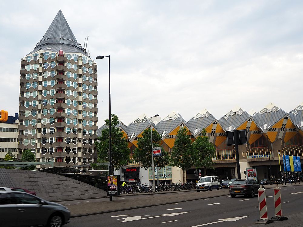 The potlood is a tall cylindrical building of about 15 stories with a round point for a roof, i.e. it's shaped like a short, fat pencil. To the right of that, the cube-shaped cube houses are visible in a row,on a sort of bridge crossing a street: yellow on one side of the cube facing down toward the street. 