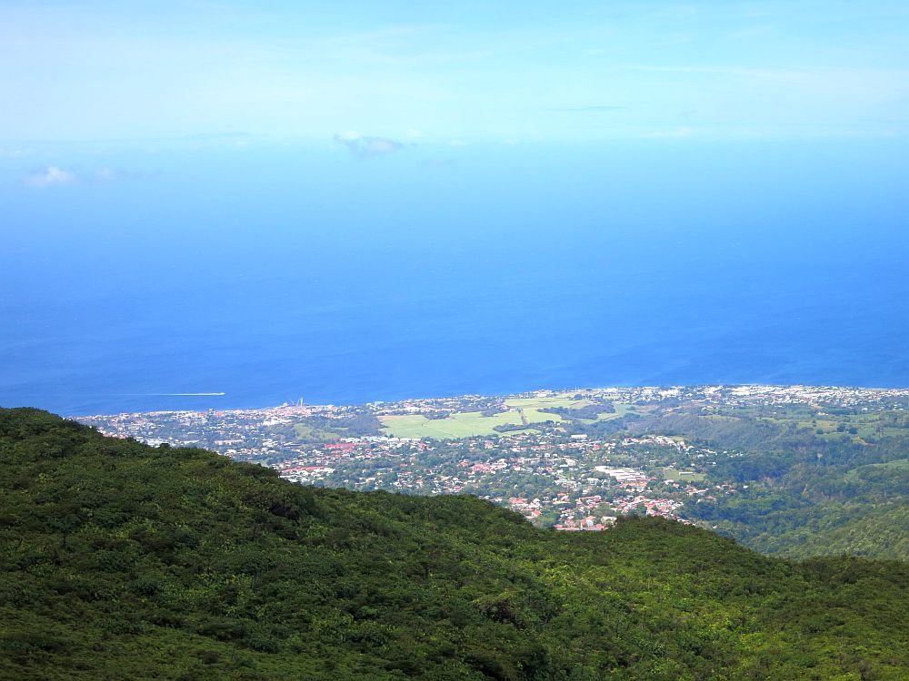 A view from the side of La Soufriere volcano in Guadeloupe shows a very green landscape, a village on the sea, and deep blue water blending into clear blue sky.