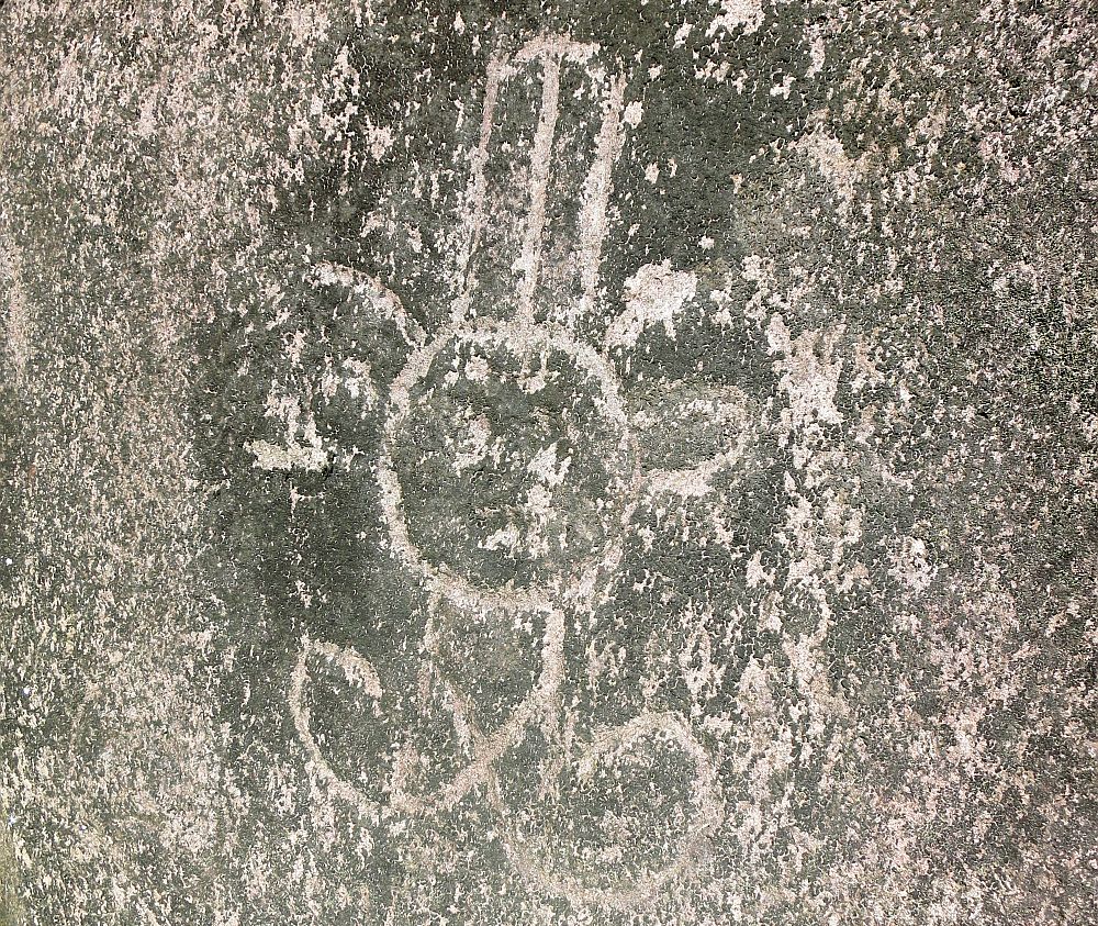 A rock carving left by Arawak people at Carved Rocks Archeological Park shows a very simple figure with a round face, a square body with large feet sticking out the bottom of the body, with no feet and no arms. the round face has what looks like large round ears and a tall hat. 