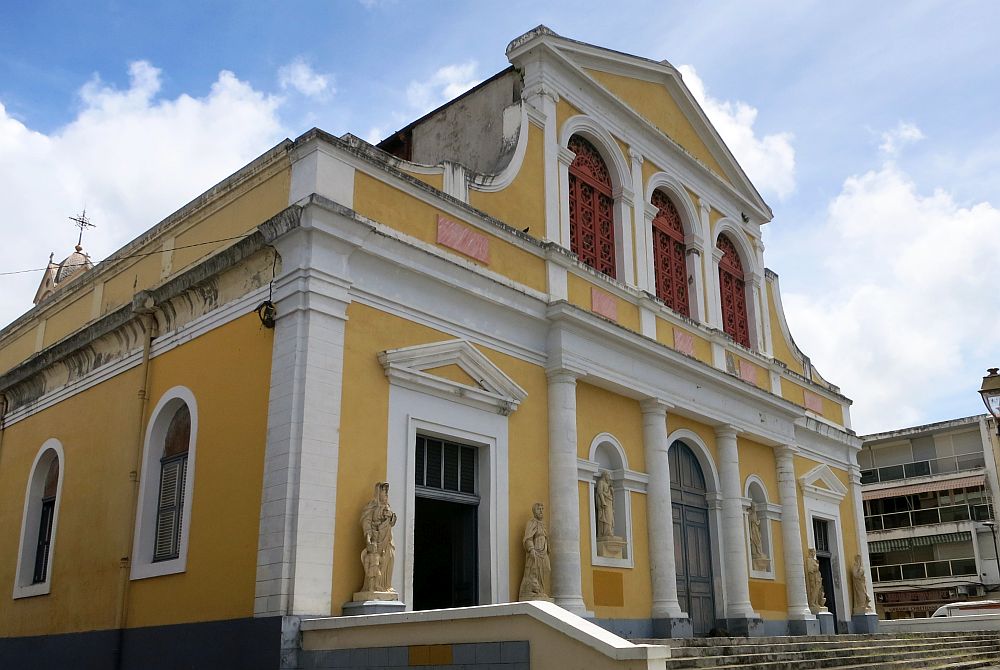 The cathedral of St. Pierre and St. Paul in Pointe-à-Pitre: The building is yellow with white edging. The ground floor has three doors, symmetrically placed; the center one has an arched top. White pillars separate them and the two statue niches between doors. The upper floor is narrower, with three arched windows and a pointed pediment. 