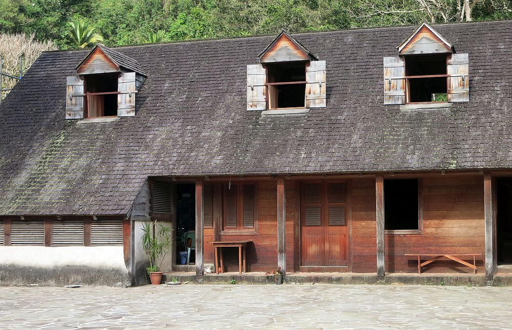 The master's house at La Grivelière coffee plantation in Guadeloupe: It has a weathered wood ground floor with a shallow porch and simple poles holding up the roof. The roof is dark brown shingles and the second floor windows stick out in dormer windows from the roof. Each dormer window has small shutters, which are open, also weathered wood.