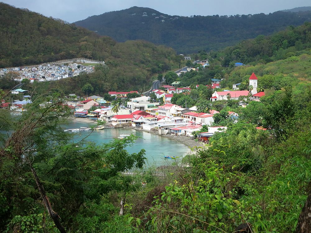 The small village is a cluster of houses facing a small bay. The houses are mostly white with red roofs. A few boats float just off the shore. A church tower with a red pointed roof sits slightly above the town up the hill that is visible behind the town. In the distance, what looks like another cluster of smaller houses is actually a cemetery.