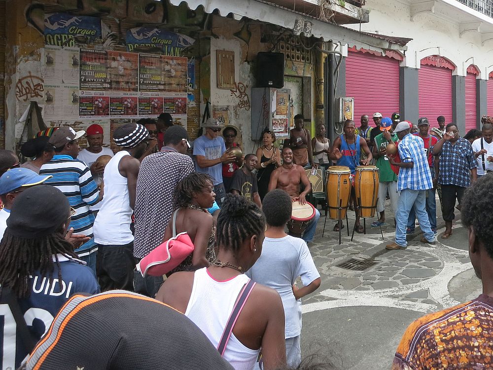 Saturday drumming session in Pointe-à-Pitre: A group of drummers, some sitting with their drums between their knees, some standing with the drums in front of them in stands. A crowd of people stand around them; many have their backs to the camera, and some of the other side of the circle of the audience is visible on the other side of the drums, facing the camera.