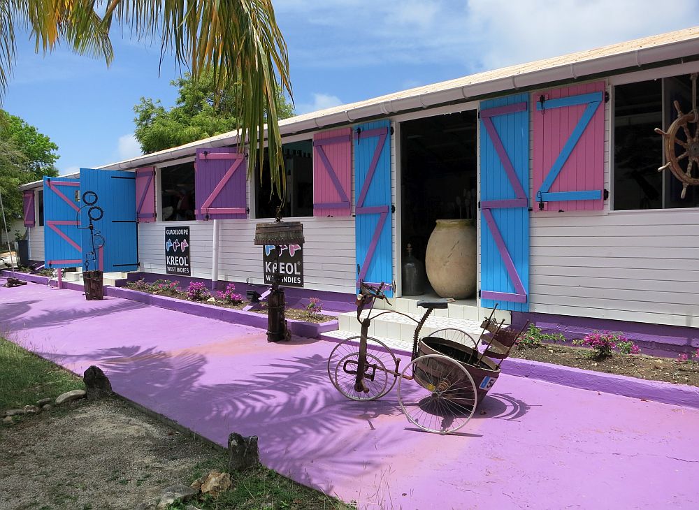 The building is a single story, quite long, with a purple-painted strip of concrete in front and shutters around the open windows (purple) and doors (blue). A few sculptures stand on the concrete strip.