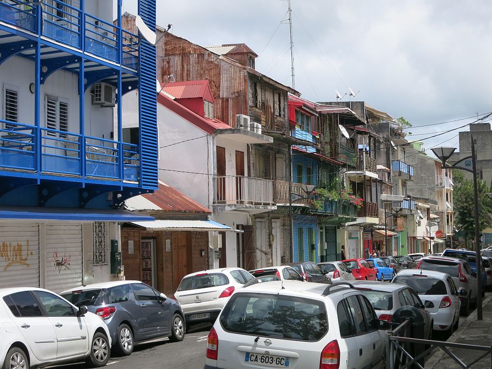 a street scene in Pointe-à-Pitre: a city street with cars parked along both sides. The other side of the street is lined with residential buildings of 3 or 4 stories each. Most have balconies and some are painted in bright colors, while some look very weatherworn.