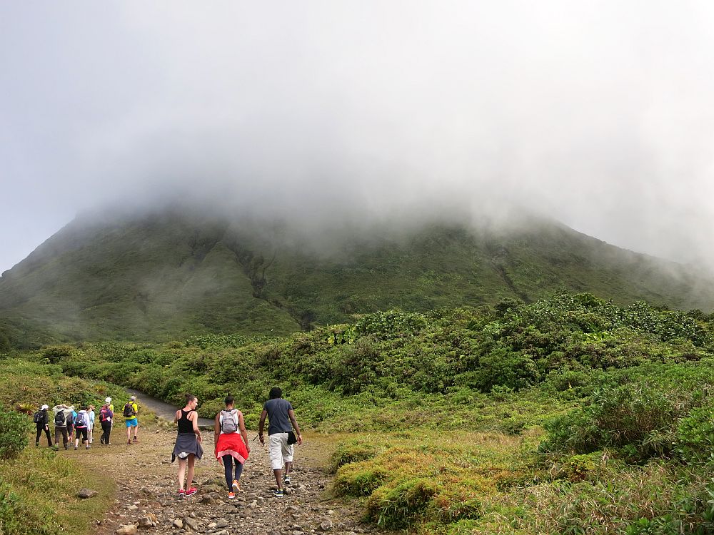 In the foreground, three people walking away from the camera on a wide dirt trail. Beyond them, a group of more people. Beyond them is the green bump of the mountain, most of which is obscured by cloud.