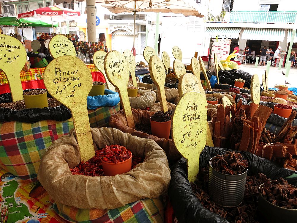 spices for sale at the market in Pointe-à-Pitre Each open pot is coverd in a plaid cloth on the sides and tucked in around the edges. Each is labeled with a large spoon-shaped yellow board sticking into the spices. Each board says the name of the spice and the price in euros.