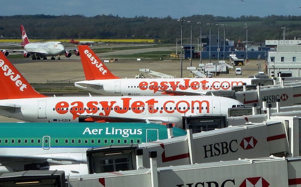 Two EasyJet planes in white and orange and an Aer LIngus plane in green are lined up at a row of gates. A larger plane in the background - a 747 - is taxiing. 