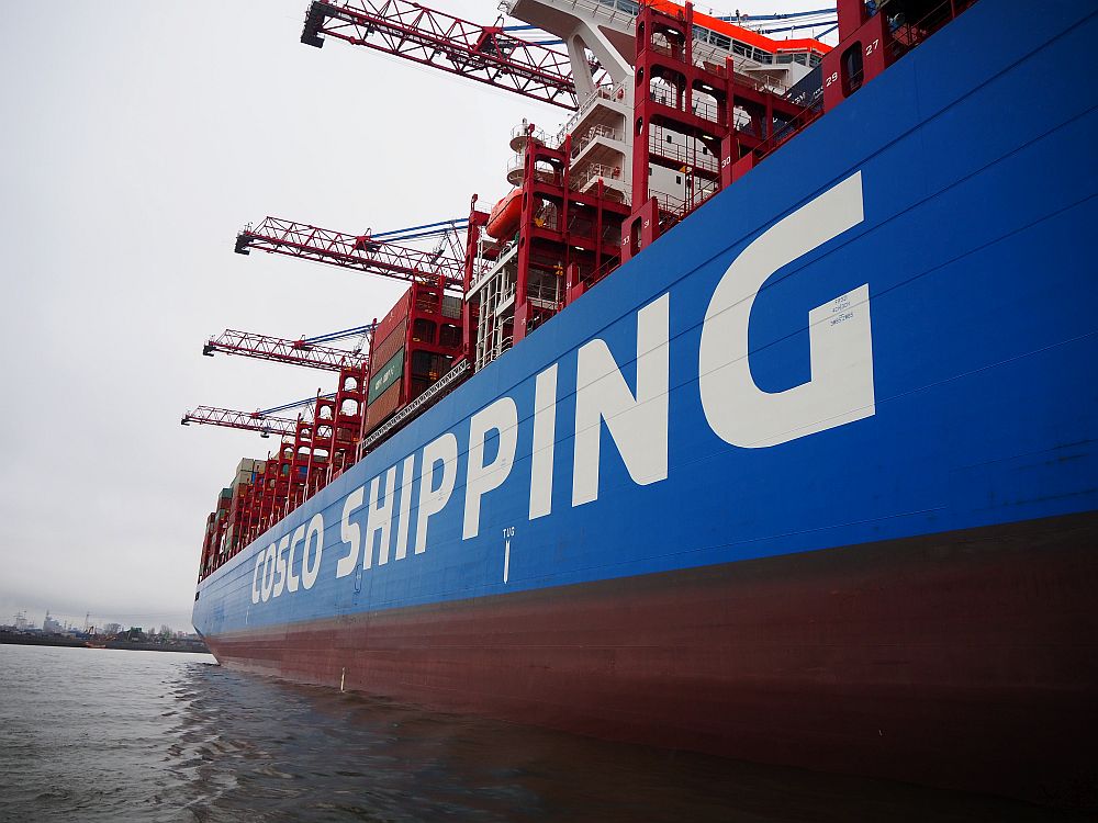 A close view of the side of a container ship, reading "Cosco Shipping" on the side, with large cranes extending over the side of the ship.