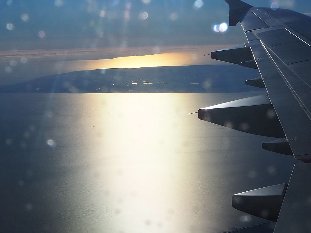 view from an airplane window, showing the sun on the water and an island in the distance, with the edge of the wing visible on the right.