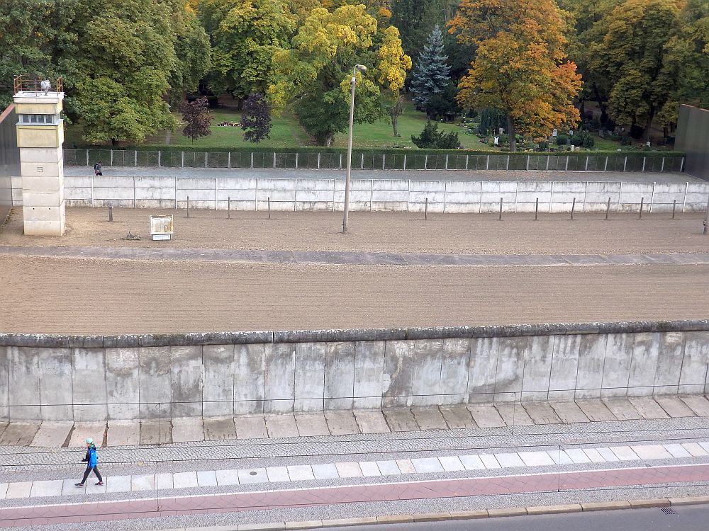 In the foreground, the concrete wall. In the background, another wall. Between is a flat, dirt-floored open area, and a guard tower stands near the far wall.