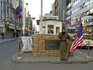 A small building in the middle of a street, with a uniformed man in front of it, holding a US flag.