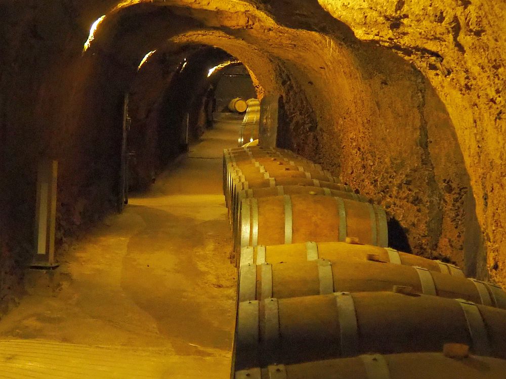 A tunnel carved from stone stretches into the distance, with a row of large barrels on their sides lining one side. 