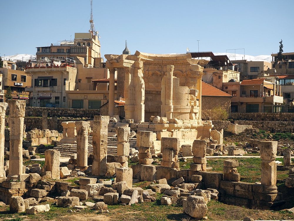 A cluster of ruins with modern buildings in the background. The largest ruin in the middle has a rounded back wall and column forming a circle. It stands on a pedestal with steps leading up to it. The ground around it has lots of pieces of stone columns, some taller, some just small stubs.