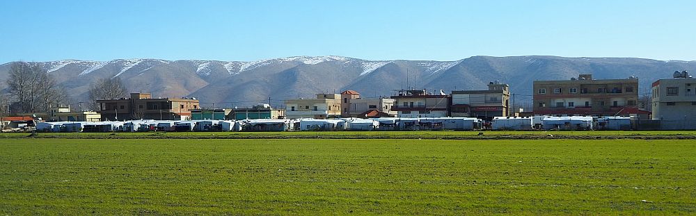 At the far side of a green field is a low line of white tents, with buildings behind them and mountains in the background.