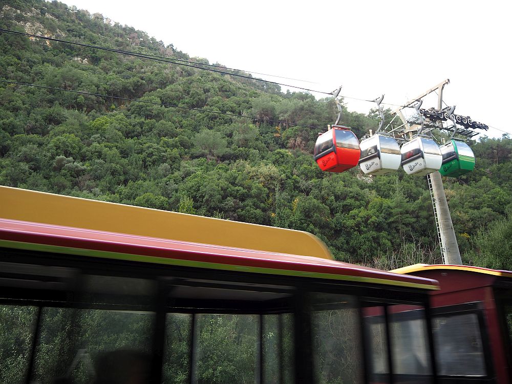 In the foreground, a blurry roof of the "train". In the background, four hanging gondolas on the ropeway cable.