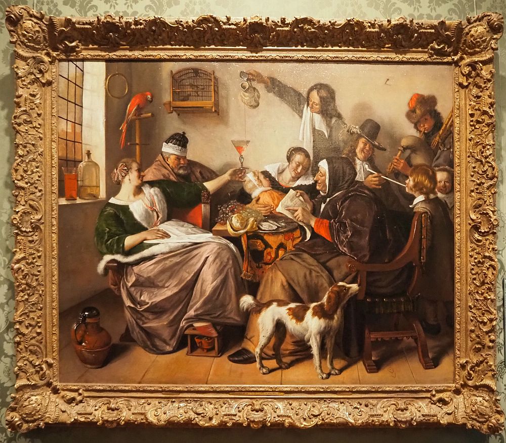 A woman reclines on the left, a pudgy man next to her. A red parrot perches above him. Behind a table, a woman holds a baby and a man reaches over her to pour wine. An old woman reads, a man gives a boy a puff from his pipe, another man plays bagpipes.