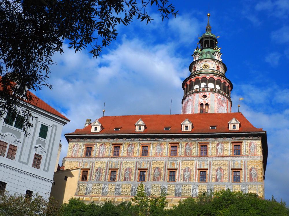 Looking up at a rectangular building and a cyclindrical tower behind it. Both are painted to look like they're made of large stone blocks with niches with statues in them.