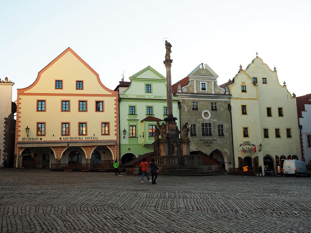 The photo shows four pastel-colored buildings on a cobbled square with a tall carved column in front.