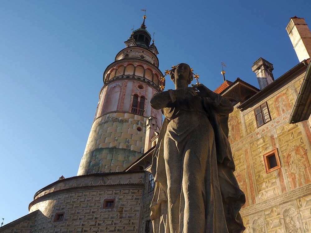 Seen from below a classical statue with a halo looms, with the conical castle tower behind it.