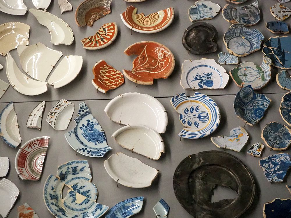 The dishes pictured are all broken and pieced together, though most also still have portions missing. Several are blue and white Delft ware, while some are plain white or earthy brown.