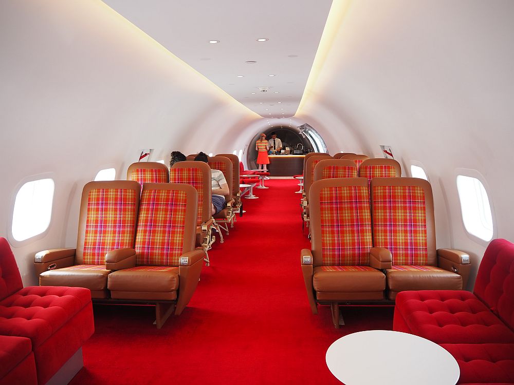 red-carpeted and white-walled, the airplane seats are red and brown leather, much more cushioned and larger than airplane seats nowadays. A bar is visible at the rear of the plane.