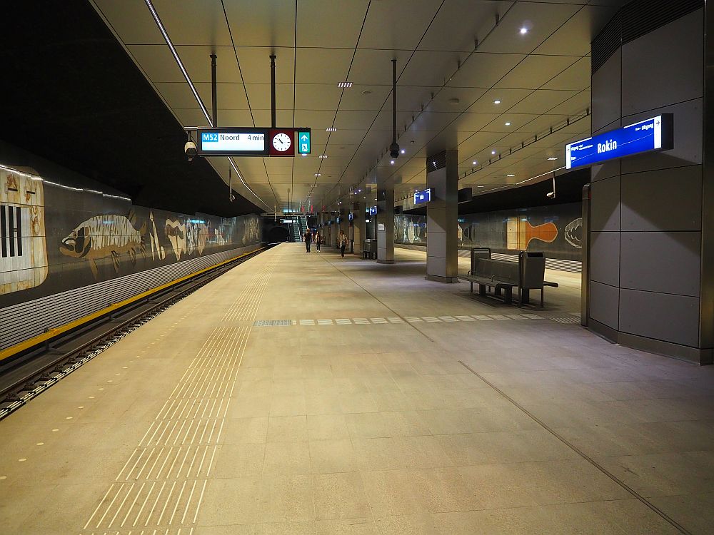 a view down the length of the station: big empty concrete platform with tracks on either side. The walls next to the tracks have large mosaics on them.