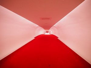 The hallway is long and upward-sloping, with white walls and ceiling and bright red carpeting on the floor.